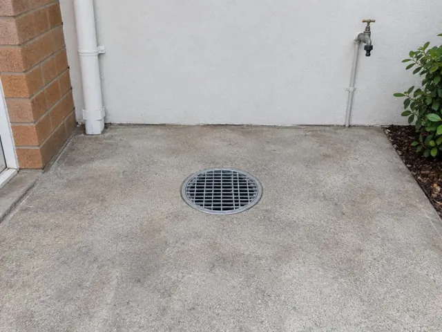 Round outdoor drain grate set in a concrete patio beside a brick wall and tap in Menai